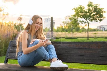Foto de Chica rubia se sienta con las piernas cruzadas en un banco y escucha música a través de auriculares en el parque al atardecer. - Imagen libre de derechos