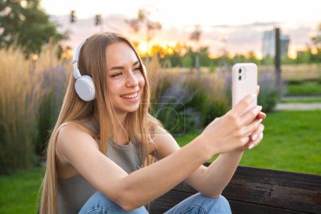 Foto de Blogger rubia dispara cuentacuentos sentado en el parque en el banco. Una joven con auriculares se toma una selfie. - Imagen libre de derechos
