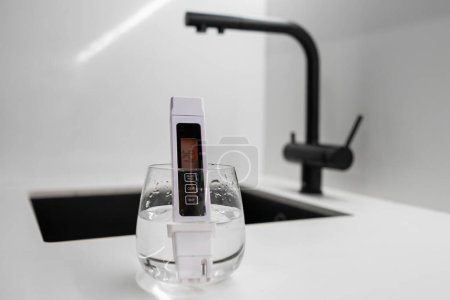 Conductometer or TDS water quality meter is immersed in a beaker of water to check purity in front of modern kitchen water tap. Dirthy water concept