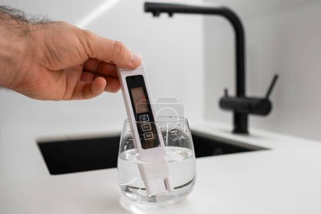 Mann Hand taucht Leitfähigkeitsmesser in ein Becherglas mit Wasser, um Reinheit vor einem modernen Küchenwasserhahn zu überprüfen. Leitfähigkeitsmesser zeigt, dass Wasser schmutzig ist 