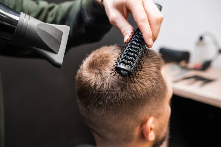 Téléchargez les photos : Brosses de coiffeur homme cheveux courts en utilisant sèche-cheveux dans le salon de coiffure. Barbier qualifié fait coiffure élégante dans le salon de beauté. Coupe de cheveux masculine - en image libre de droit