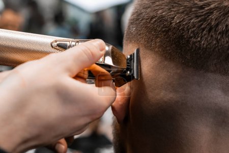 Téléchargez les photos : Coiffeur rase les cheveux du client avec une tondeuse électrique dans le gros plan moderne du salon de coiffure. Barber fait coiffure élégante contour dans le salon de beauté - en image libre de droit