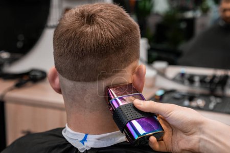 Friseur entfernt Haare hinter dem Ohr des Kunden und Okziput mit Rasierer in Friseur-Nahaufnahme. Friseur nutzt moderne Maschine für stilvollen Haarschnitt im Salon