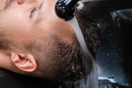 Téléchargez les photos : Coiffeur lave les cheveux de l'homme de shampooing avec de l'eau chaude dans le salon de coiffure gros plan. Barber prend soin des cheveux du client masculin dans le salon professionnel - en image libre de droit