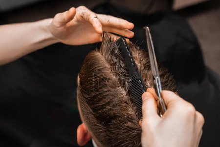 Stylist cuts male client hair using comb and scissors in barbershop closeup. Barber with professional tools serves man in salon