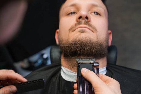 Téléchargez les photos : Maître coupe la barbe du client avec tondeuse automatique dans le gros plan du salon de coiffure. Coiffeur fait la coupe de cheveux avec un équipement moderne dans le salon de toilettage - en image libre de droit