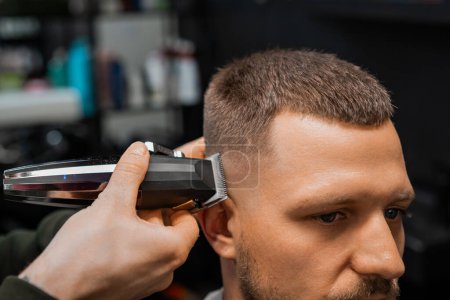Téléchargez les photos : Barbier qualifié utilise tondeuse automatique pour couper les cheveux du client sur le temple dans le salon de coiffure gros plan. Coiffeur fait coiffure élégante par miroir salon - en image libre de droit