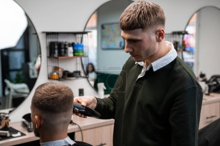 Téléchargez les photos : Barber utilise une machine à raser pour couper les cheveux du jeune homme dans le gros plan du salon de coiffure. Maître fait la coupe de cheveux moderne au client masculin dans le salon professionnel - en image libre de droit