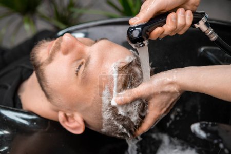 Téléchargez les photos : Coiffeur qualifié rince tendrement les cheveux d'un client de shampooing à l'eau chaude. - en image libre de droit