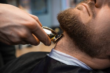 Téléchargez les photos : Styliste coupe les cheveux sur le cou créant une forme de barbe élégante pour l'homme avec tondeuse dans le gros plan de salon de coiffure. Barber sert client masculin dans le salon de beauté - en image libre de droit