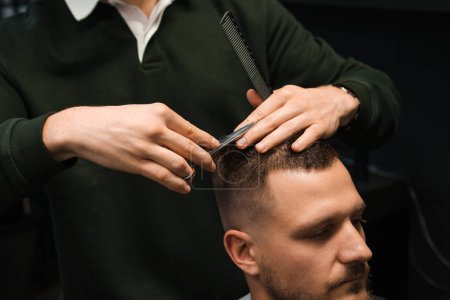 Man hairstylist cutting the clients brunette hair with scissors at the barbershop