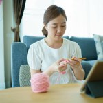 Happy cheerful Asian woman doing a crochet in living room in free time. Skillful Asian craftswoman knitting a handcraft crochet sweater and hat in cozy living room.