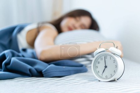 Foto de Mujer joven asiática durmiendo en la acogedora cama por la mañana temprano y tratando de apagar o dormir un reloj despertador. Mujer somnolienta perezosa se despierta tarde en la mañana. - Imagen libre de derechos