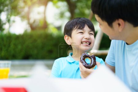Foto de Feliz alegre padre asiático e hijo comiendo una rosquilla juntos durante el picnic en el jardín. Emocionado niño encantador comiendo una rosquilla con su padre. Concepto de actividad familiar fin de semana. - Imagen libre de derechos