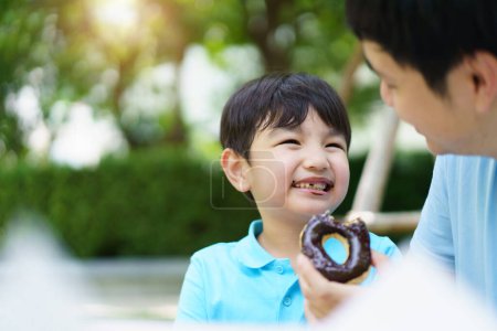 Foto de Feliz alegre padre asiático e hijo comiendo una rosquilla juntos durante el picnic en el jardín. Emocionado niño encantador comiendo una rosquilla con su padre. Concepto de actividad familiar fin de semana. - Imagen libre de derechos