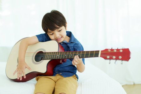 Happy cheerful Asian little young boy practices playing an acoustic guitar in bedroom. Lovely Asian little boy playing a guitar.