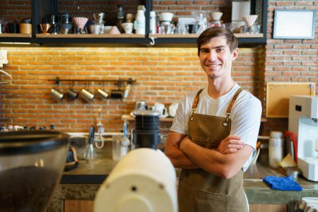 Photo for Professional young coffee barista standing inside the counter bar in the coffee shop and making arms crossed and looking at camera, handsome - good looking caucasian ethnic barista posing. - Royalty Free Image