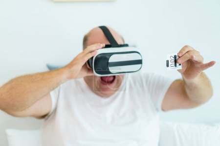 Foto de Caucásico hombre gordo de gran tamaño mostrando un condón en su mano mientras ve un entretenimiento en las gafas o gafas de realidad virtual. - Imagen libre de derechos