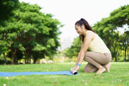 Photo for Happy beautiful Asian woman rolling out an exercise - yoga mat on the grass field to prepare doing a yoga and mediation exercise. Woman preparing a yoga or body weight and meditating exercise. - Royalty Free Image
