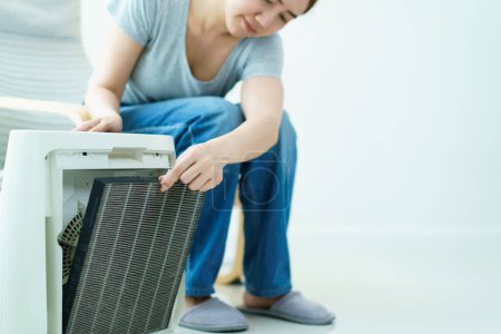 Asian middle adult woman changing a dirty hepa filter in the portable air purifier machine. Woman replaces a dirty and dusty air purifier filter in living room. Air pollution pm 2.5 prevention concept.