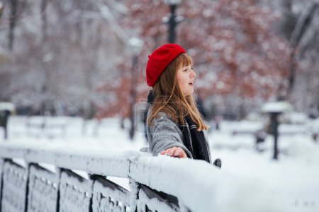 Foto de Joven pelirroja en rojo sombrero de invierno caminando parque nevado - Imagen libre de derechos