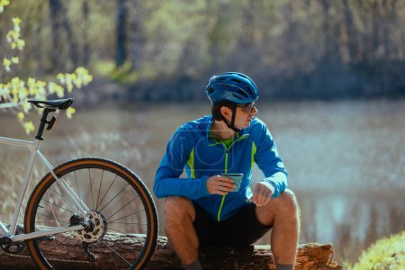 Téléchargez les photos : Jeune homme assis sur le rondin avec vélo et profiter de son temps le matin dans la nature et boire du thé - en image libre de droit