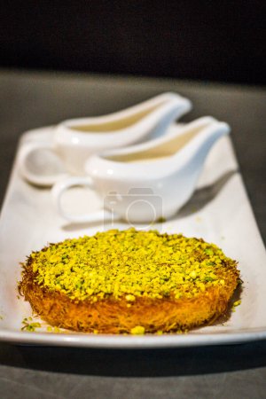 Téléchargez les photos : Un Dessert de Kunafa servi avec des pistaches écrasées sur le dessus et de la crème et du sirop de sucre sur le côté - en image libre de droit
