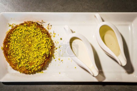 Téléchargez les photos : Un Dessert de Kunafa servi avec des pistaches écrasées sur le dessus et de la crème et du sirop de sucre sur le côté - en image libre de droit