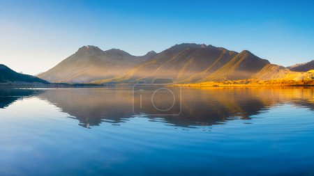 Téléchargez les photos : Lake and mountains in a valley at dawn. Reflections on the surface of the lake. Mountain landscape at sunrise. Foggy morning. Natural landscape with bright sunshine. - en image libre de droit