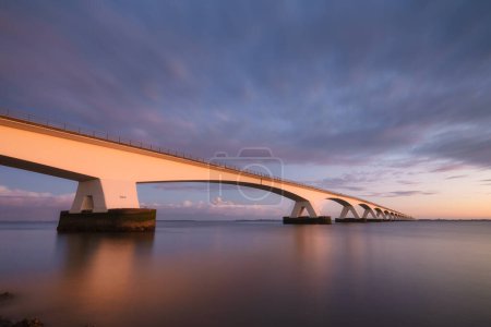 Téléchargez les photos : Pont Zeeland, Pays-Bas. Un long pont sur la mer au coucher du soleil. Photo longue exposition. Paysage lors d'un coucher de soleil lumineux. La mer et le pont. - en image libre de droit