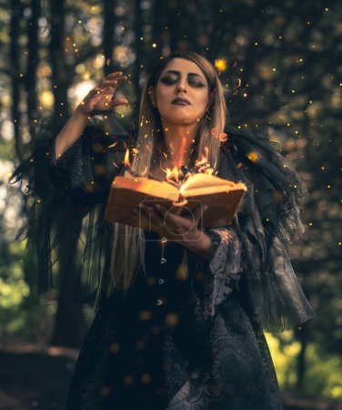 Foto de Bruja cautivadora en un misterioso bosque de Halloween, conjurando magia con un libro de hechizos en llamas en una atmósfera espeluznante - Imagen libre de derechos