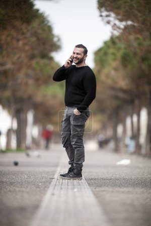 Photo for Natural looking guy posing outdoors, wearing casual clothes, holding a smartphone and talking,  no post edit - Royalty Free Image