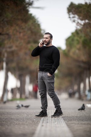 Photo for Natural looking guy posing outdoors, wearing casual clothes, holding a smartphone and talking,  no post edit - Royalty Free Image