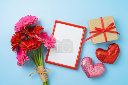 Téléchargez les photos : Cadre photo Saint-Valentin maquette avec bouquet de fleurs de marguerite gerbera, boîte cadeau et formes de coeur sur fond bleu. Vue de dessus, plan plat - en image libre de droit