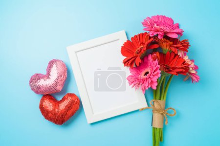 Photo for Valentine's day white picture frame mock up with gerbera daisy flower bouquet and heart shapes  on blue background. Top view, flat lay - Royalty Free Image