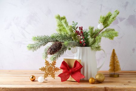 Foto de Decoración navideña con ramas de pino en jarrón, caja de regalo y adornos dorados sobre mesa de madera sobre fondo brillante - Imagen libre de derechos