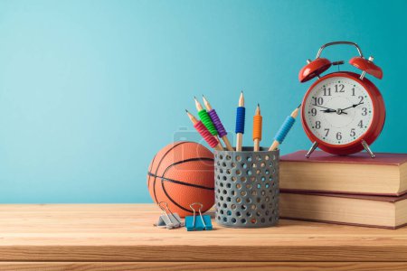 Téléchargez les photos : Concept de retour à l'école avec crayons, ballon de basket, livres et réveil sur table en bois sur fond bleu - en image libre de droit