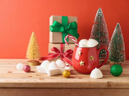 Foto de Copa de chocolate caliente de Navidad con malvavisco, caja de regalo y decoraciones en mesa de madera sobre fondo rojo - Imagen libre de derechos