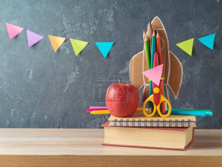Foto de Concepto de regreso a la escuela con cohete, lápices, manzana y libro sobre mesa de madera sobre fondo pizarra - Imagen libre de derechos