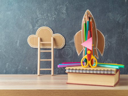 Foto de Concepto de regreso a la escuela con cohete, lápices y libro sobre mesa de madera sobre fondo pizarra - Imagen libre de derechos