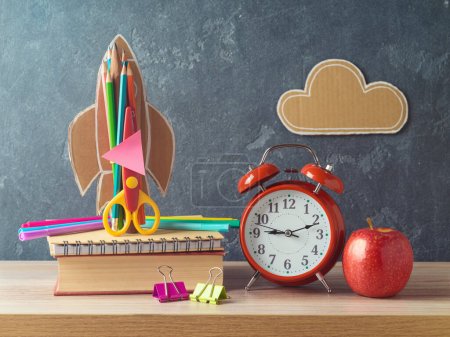 Téléchargez les photos : Concept de retour à l'école avec fusée, crayons, pomme, réveil et livre sur table en bois sur fond de tableau noir - en image libre de droit