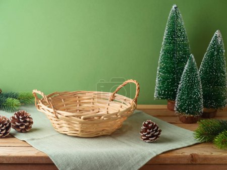 Foto de Cesta vacía con mantel sobre mesa de madera sobre pared verde y fondo decorativo de pino. Maqueta de Navidad para el diseño y la exhibición del producto. - Imagen libre de derechos