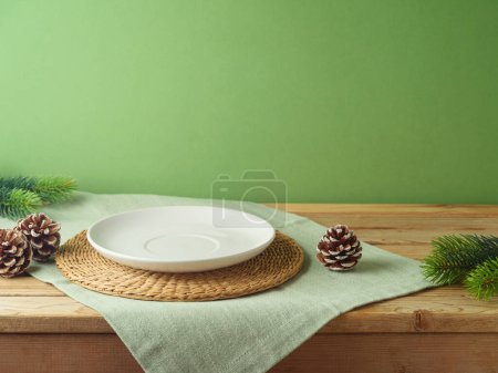 Foto de Placa blanca vacía con mantel sobre mesa de madera sobre pared verde y fondo decorativo de pino. Maqueta de Navidad para el diseño y la exhibición del producto. - Imagen libre de derechos