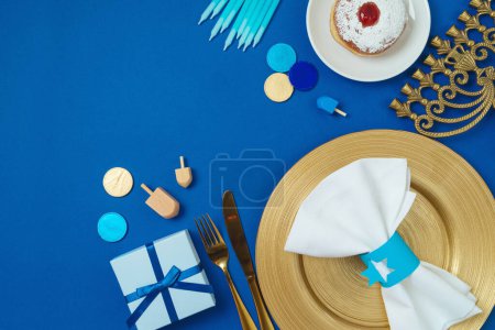 Foto de Judío concepto de Hanukkah vacaciones con plato de oro, donas tradicionales, menorah y caja de regalo sobre fondo azul. Vista superior, plano - Imagen libre de derechos