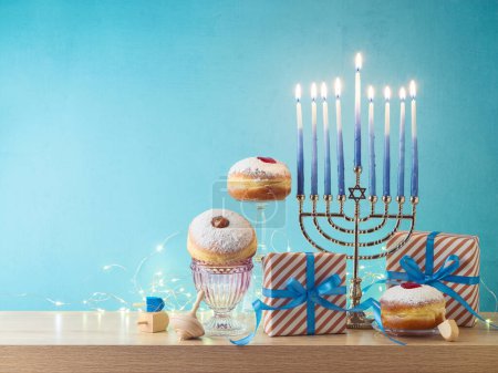 Photo for Jewish holiday Hanukkah creative composition with menorah, traditional donuts and gift box on wooden table over blue background - Royalty Free Image