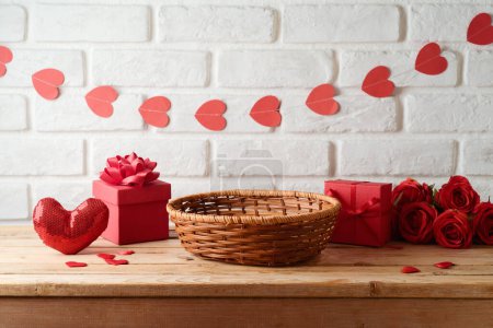Foto de Cesta vacía en mesa de madera con caja de regalo y guirnalda en forma de corazón sobre fondo de pared de ladrillo. Burla del día de San Valentín para el diseño y la exhibición del producto - Imagen libre de derechos