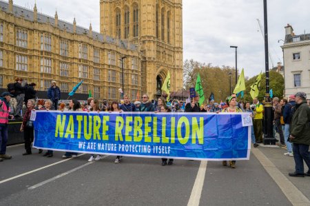 Foto de LONDRES - 22 de abril de 2023: Manifestantes de la XR presencian la marcha, llevando la bandera de la Rebelión de la Naturaleza frente a las Casas del Parlamento en Londres, uniéndose por la justicia ambiental y social. - Imagen libre de derechos