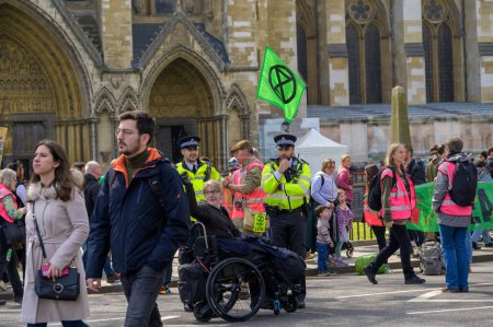 Foto de LONDRES - 22 de abril de 2023: Diálogo inclusivo: Oficiales de la Policía Metropolitana se relacionan con un usuario de silla de ruedas en medio de los manifestantes del XR en la marcha de la rebelión contra la extinción en Londres. - Imagen libre de derechos