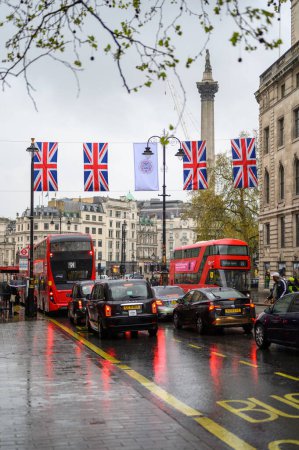 Foto de LONDRES - 24 de abril de 2023: Una escena por excelencia en Londres: taxis negros clásicos, autobuses rojos de dos pisos, banderas Union Jack y el telón de fondo histórico de Trafalgar Square. - Imagen libre de derechos