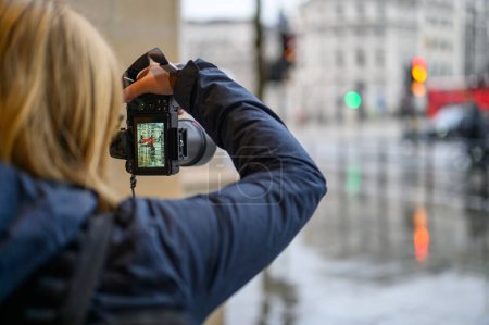 Foto de LONDRES - 24 de abril de 2023: Una fotógrafa rubia captura la esencia de Londres en un día lluvioso, centrándose en un autobús rojo de dos pisos. - Imagen libre de derechos
